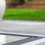 Thumbnail of http://A%20close-up%20view%20of%20a%20modern%20outdoor%20patio%20with%20grey%20tiles,%20a%20reflective%20surface,%20green%20grass,%20a%20large%20planter,%20and%20a%20wooden%20fence%20in%20the%20background.