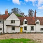 Thumbnail of http://A%20white,%20two-storey%20cottage%20with%20a%20red%20tiled%20roof,%20multiple%20chimneys,%20and%20a%20bright%20yellow%20front%20door,%20set%20on%20a%20gravel%20drive%20with%20lawn%20and%20trees%20in%20the%20background.