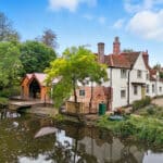 Thumbnail of http://Historic%20white%20cottage%20with%20brick%20chimneys%20and%20a%20wooden%20extension%20sits%20beside%20a%20small%20pond,%20surrounded%20by%20trees%20and%20greenery%20under%20a%20partly%20cloudy%20sky.