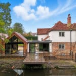 Thumbnail of http://A%20riverside%20house%20with%20a%20mix%20of%20modern%20and%20traditional%20architecture,%20featuring%20large%20glass%20doors,%20wooden%20decking,%20brick%20walls,%20and%20a%20red-tiled%20roof,%20surrounded%20by%20trees.