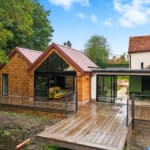 Thumbnail of http://Modern%20wooden%20house%20extension%20with%20glass%20walls%20and%20a%20red%20roof,%20connected%20to%20an%20older%20brick%20home,%20features%20a%20spacious%20decking%20surrounded%20by%20greenery.