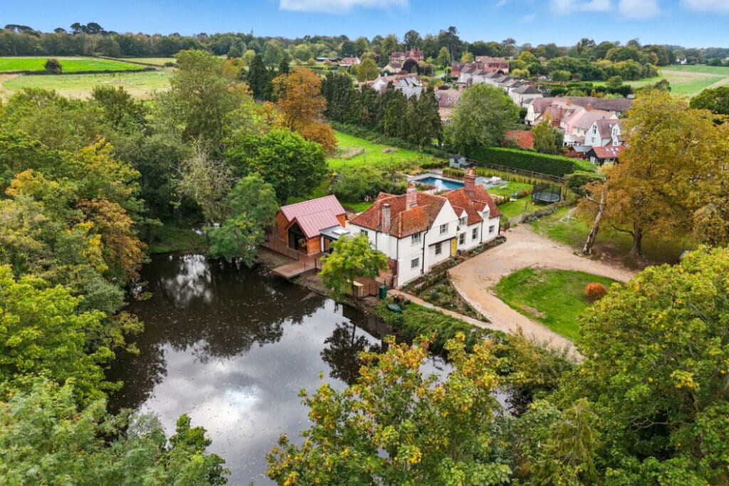 A white house with red roofs sits beside a pond, surrounded by trees and greenery, with additional houses and fields in the background.