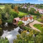 Thumbnail of http://A%20white%20house%20with%20red%20roofs%20sits%20beside%20a%20pond,%20surrounded%20by%20trees%20and%20greenery,%20with%20additional%20houses%20and%20fields%20in%20the%20background.