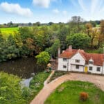 Thumbnail of http://Aerial%20view%20of%20a%20white%20country%20house%20with%20a%20red-tiled%20roof%20beside%20a%20pond,%20surrounded%20by%20fields%20and%20trees%20under%20a%20partly%20cloudy%20sky.