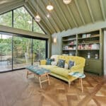 Thumbnail of http://A%20modern%20living%20room%20with%20vaulted%20green%20ceiling,%20large%20sliding%20glass%20doors,%20yellow%20sofa,%20built-in%20bookcase,%20and%20herringbone%20wood%20flooring,%20overlooking%20a%20deck%20and%20greenery%20outside.