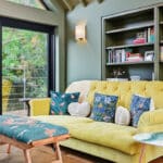 Thumbnail of http://A%20living%20room%20with%20a%20yellow%20sofa,%20patterned%20cushions,%20a%20small%20stool,%20a%20round%20side%20table,%20built-in%20bookcases,%20and%20large%20glass%20doors%20overlooking%20greenery.