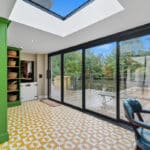Thumbnail of http://Bright%20room%20with%20patterned%20yellow%20floor%20tiles,%20green%20cupboards,%20and%20glass%20doors%20opening%20to%20a%20wooden%20decking%20with%20trees%20and%20blue%20sky%20visible%20outside.