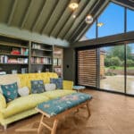 Thumbnail of http://A%20modern%20living%20room%20with%20a%20yellow%20sofa,%20built-in%20bookcase,%20and%20large%20floor-to-ceiling%20windows%20overlooking%20a%20patio%20and%20garden.%20The%20ceiling%20is%20vaulted%20with%20exposed%20beams.