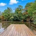 Thumbnail of http://Wooden%20decking%20with%20metal%20railings%20extends%20over%20a%20calm%20river,%20surrounded%20by%20trees%20with%20green%20and%20some%20autumn-coloured%20leaves%20under%20a%20blue%20sky%20with%20clouds.