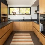 Thumbnail of http://Modern%20kitchen%20with%20light%20wood%20cupboards,%20black%20worktops,%20a%20striped%20rug,%20window%20above%20the%20sink,%20and%20decorative%20items%20on%20shelves%20and%20worktops.