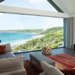 Thumbnail of http://A%20modern%20living%20room%20with%20large%20windows%20offers%20a%20panoramic%20view%20of%20a%20coastal%20landscape%20with%20blue%20water,%20sandy%20beach,%20and%20green%20hills.