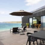 Thumbnail of http://A%20modern%20seaside%20patio%20with%20black%20furniture,%20a%20large%20parasol,%20and%20glass%20balustrade%20overlooks%20a%20calm%20blue%20sea%20and%20distant%20cliffs%20on%20a%20sunny%20day.