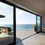 Thumbnail of http://Modern%20living%20room%20with%20large%20glass%20doors%20opening%20to%20a%20patio%20with%20chairs,%20parasol,%20and%20sea%20view%20under%20a%20clear%20sky.