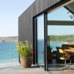 Thumbnail of http://Modern%20seaside%20home%20with%20large%20glass%20doors%20opening%20to%20a%20patio,%20overlooking%20a%20coastal%20landscape%20with%20cliffs%20and%20sea,%20and%20a%20dining%20table%20visible%20inside.