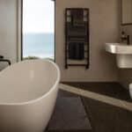 Thumbnail of http://Modern%20bathroom%20with%20a%20freestanding%20bath,%20wall-mounted%20basin,%20heated%20towel%20rail%20with%20towels,%20toilet,%20and%20window%20showing%20a%20sea%20view.
