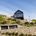 Thumbnail of http://Modern%20house%20with%20large%20glass%20windows%20and%20angular%20roof%20sits%20on%20a%20hillside,%20surrounded%20by%20stone%20walls,%20greenery,%20and%20clear%20blue%20sky.