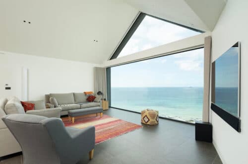 Modern living room with grey sofas, a red rug, and a large window offering a clear view of the sea and sky. A flat-screen TV is mounted on the right wall.