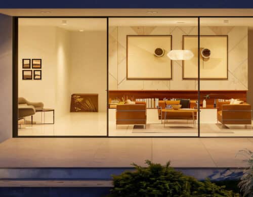 View through large glass windows showing a modern, well-lit living room with contemporary furniture, wall art, and built-in shelves, surrounded by a landscaped garden.