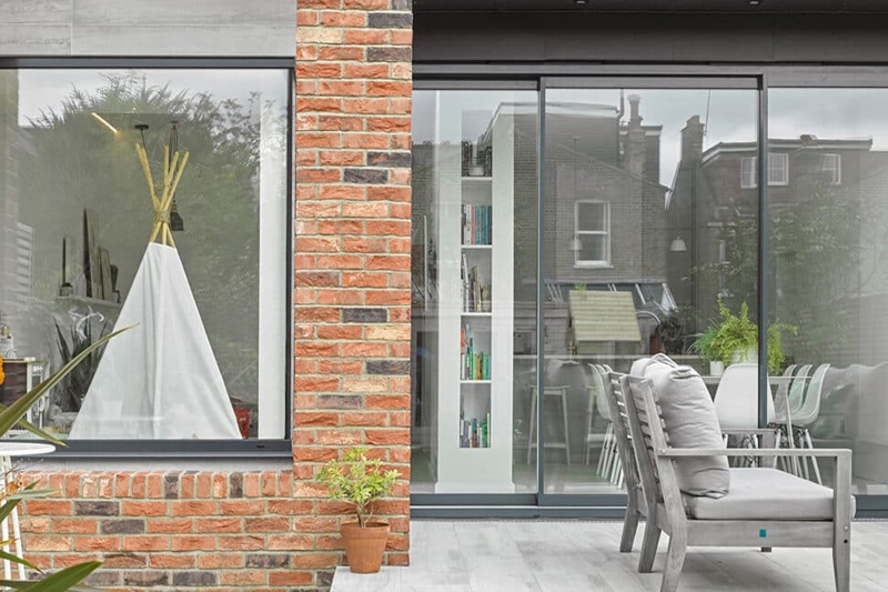 Merstham glass bifold doors with red brick chimney, opening onto contemporary patio with seating