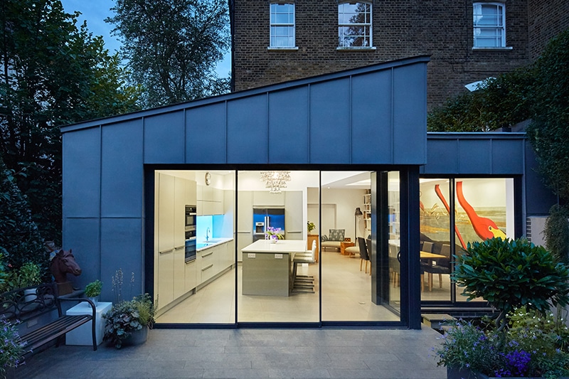 Modern blue-clad extension with large glazed bifold doors opening to a kitchen and living space, featuring black framing and floor-to-ceiling glazing.