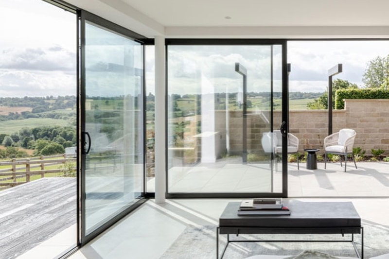 Modern bifold doors with black frames opening onto a sunlit terrace overlooking countryside landscape.