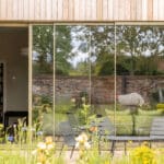 Thumbnail of http://Bifold%20doors%20opening%20from%20timber-clad%20eco-home%20onto%20garden%20with%20patio%20seating%20and%20stone%20wall%20backdrop.