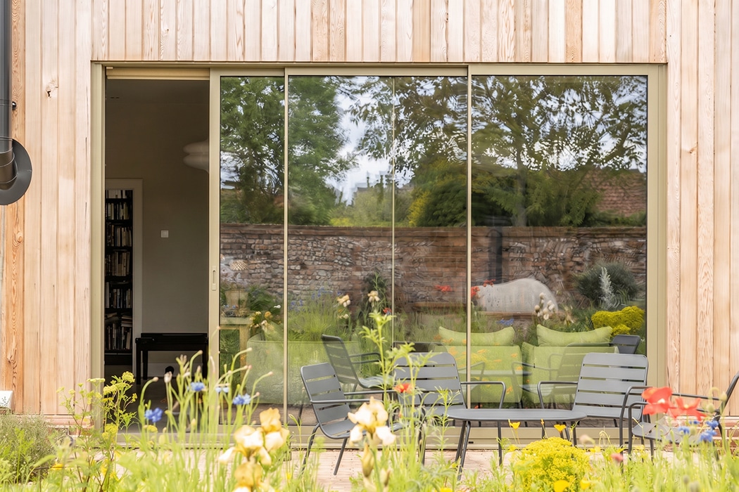 Bifold doors opening from timber-clad eco-home onto garden with patio seating and stone wall backdrop.