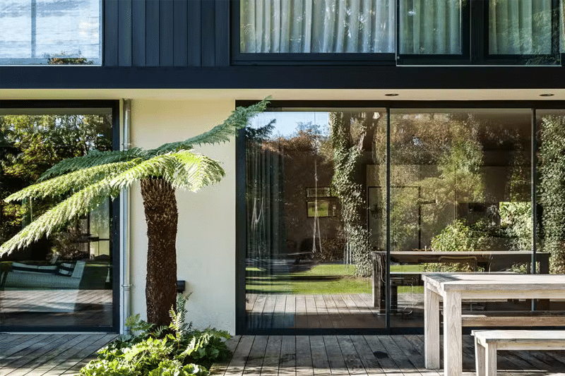 Modern bifold doors opening onto a timber deck with garden views and ivy-covered trees beyond.