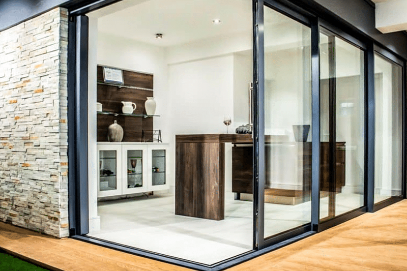 Sunflex UK bifold doors with black frames opening into modern showroom display with wooden cabinetry and stone feature wall.