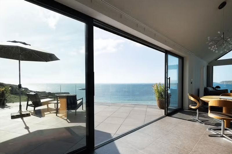 Large bifold sliding doors opening onto a coastal terrace with sea views, demonstrating contemporary glazing design.