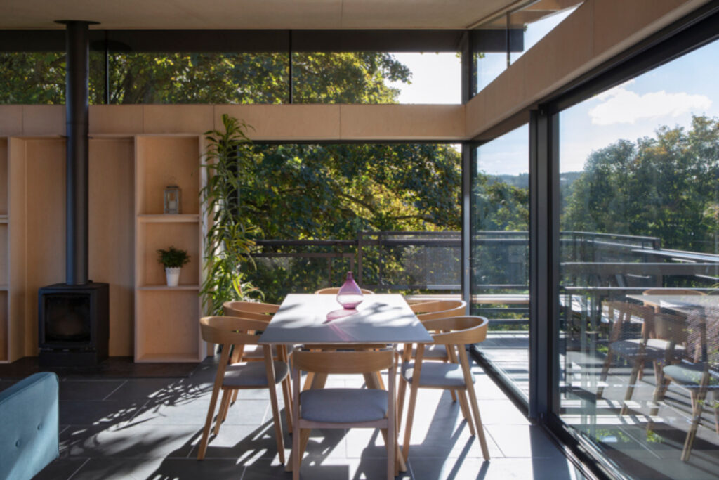 Modern dining area with bifold doors opening onto a terrace overlooking woodland landscape, wooden furniture and natural light.